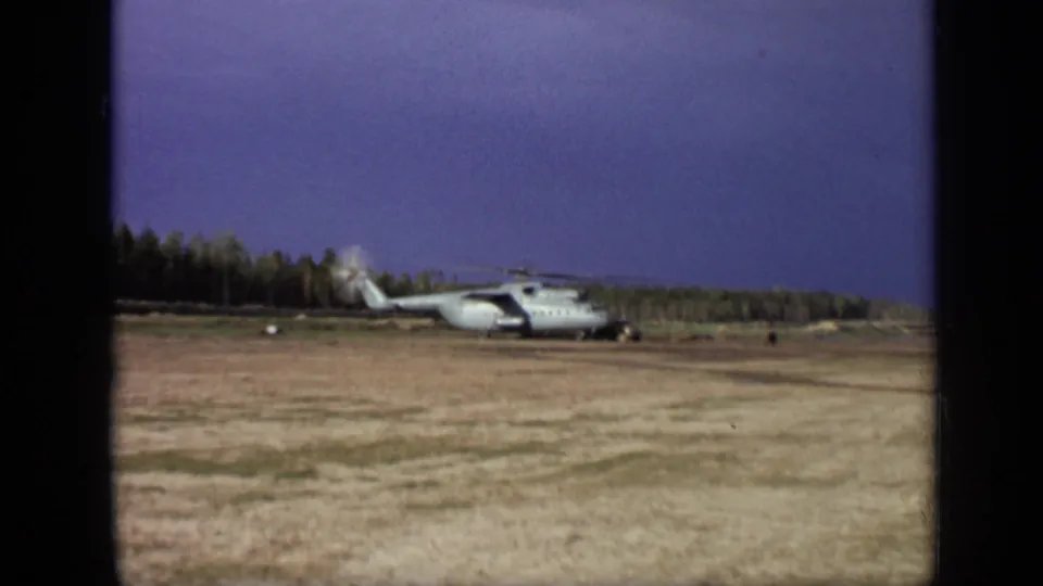 Wider 1971 Moscow 8mm view showing the parked helicopter at a distance on an open field with trees behind it.