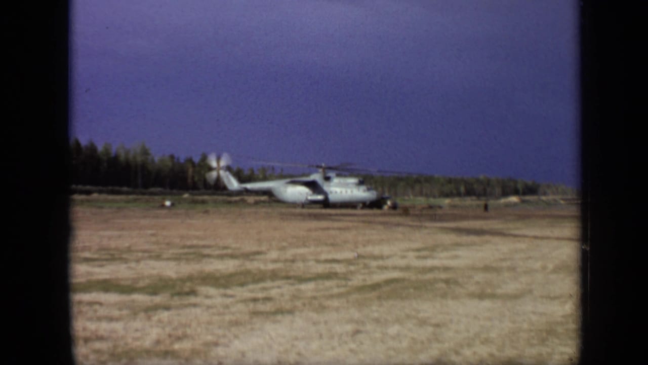1971 8mm frame near Moscow showing a large landed helicopter in a dry open field with spinning rotors and a tree line beyond.