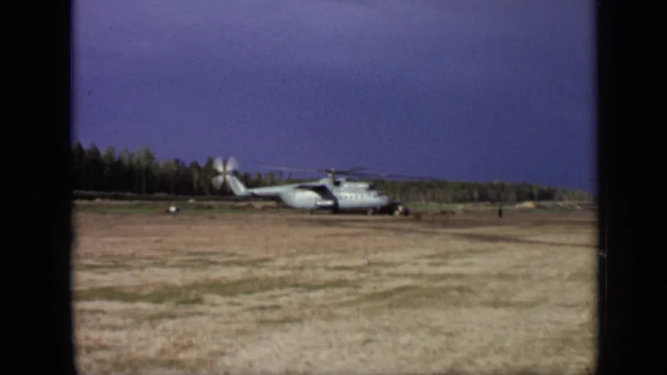 1971 8mm frame near Moscow showing a large landed helicopter in a dry open field with spinning rotors and a tree line beyond.