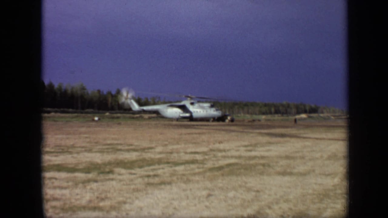 Distant helicopter on a field with spinning rotors, trees, and open sky.