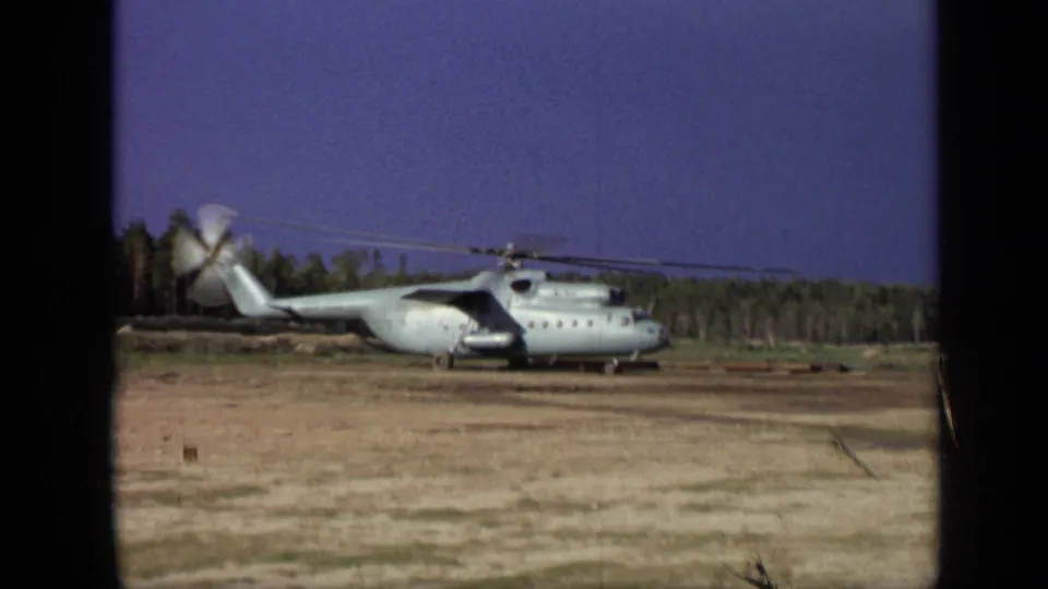 1971 8mm frame near Moscow showing a closer side view of the landed helicopter with rotating blades and evergreen trees behind it.