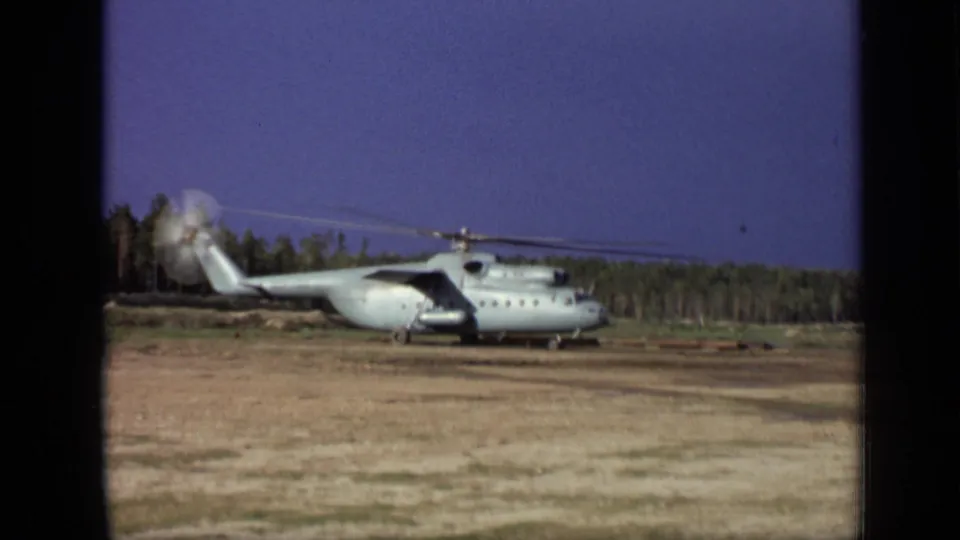 1971 8mm frame near Moscow showing the landed helicopter in profile, with rotor motion blur over a dry field and wooded backdrop.