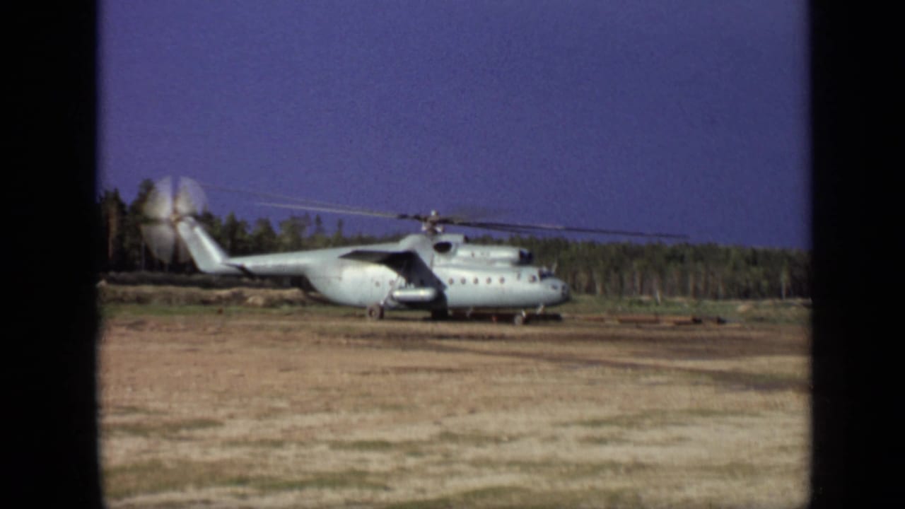 Large helicopter parked on a barren field with its rotor spinning near a tree line.