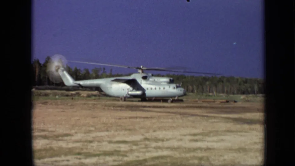 8mm footage frame of a large helicopter viewed from the side on a dry field near Moscow, with spinning rotor blur and a dark tree line.