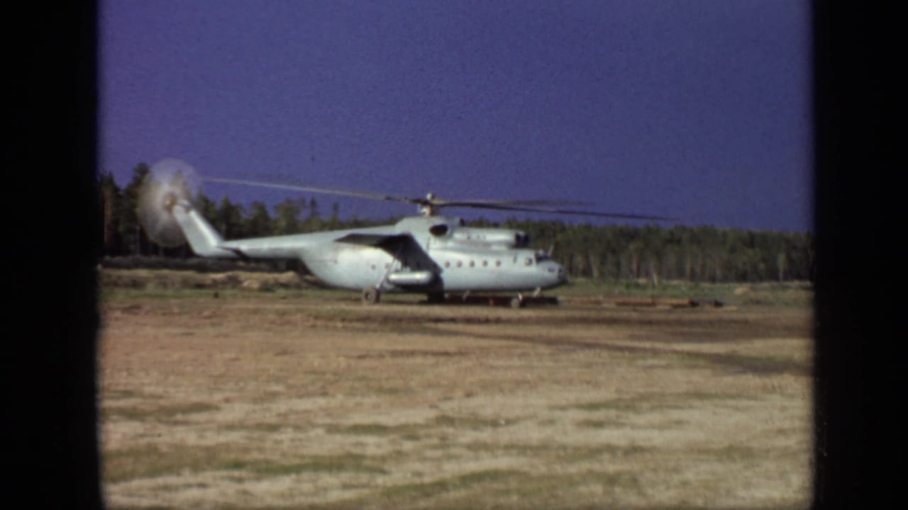 1971 Moscow 8mm frame showing a large helicopter stationary on a dirt field with blurred spinning rotors and trees behind it.
