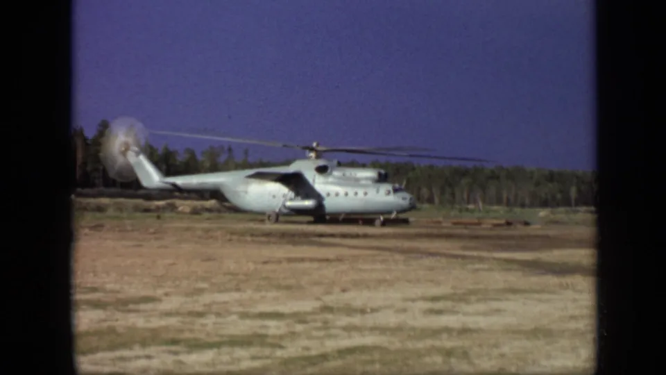 1971 Moscow 8mm frame showing a large helicopter stationary on a dirt field with blurred spinning rotors and trees behind it.