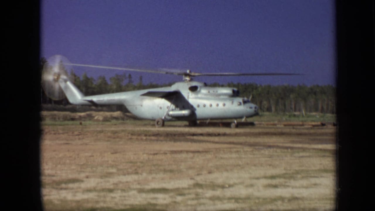Large helicopter with spinning rotors seen broadside on a dusty open field.