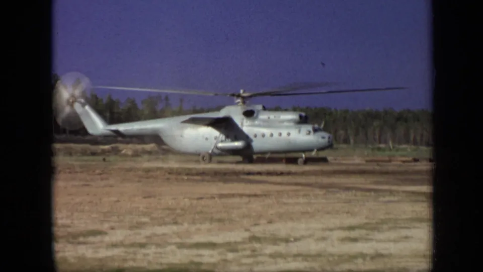 1971 Moscow 8mm frame showing the helicopter from a steady distance, with rotor blur, dirt field, and treeline visible.