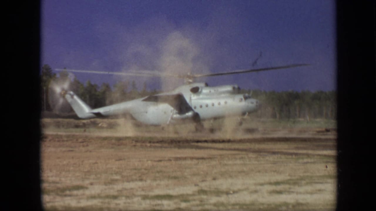 Light-colored helicopter on a dirt field beginning takeoff as rotor wash raises dust.