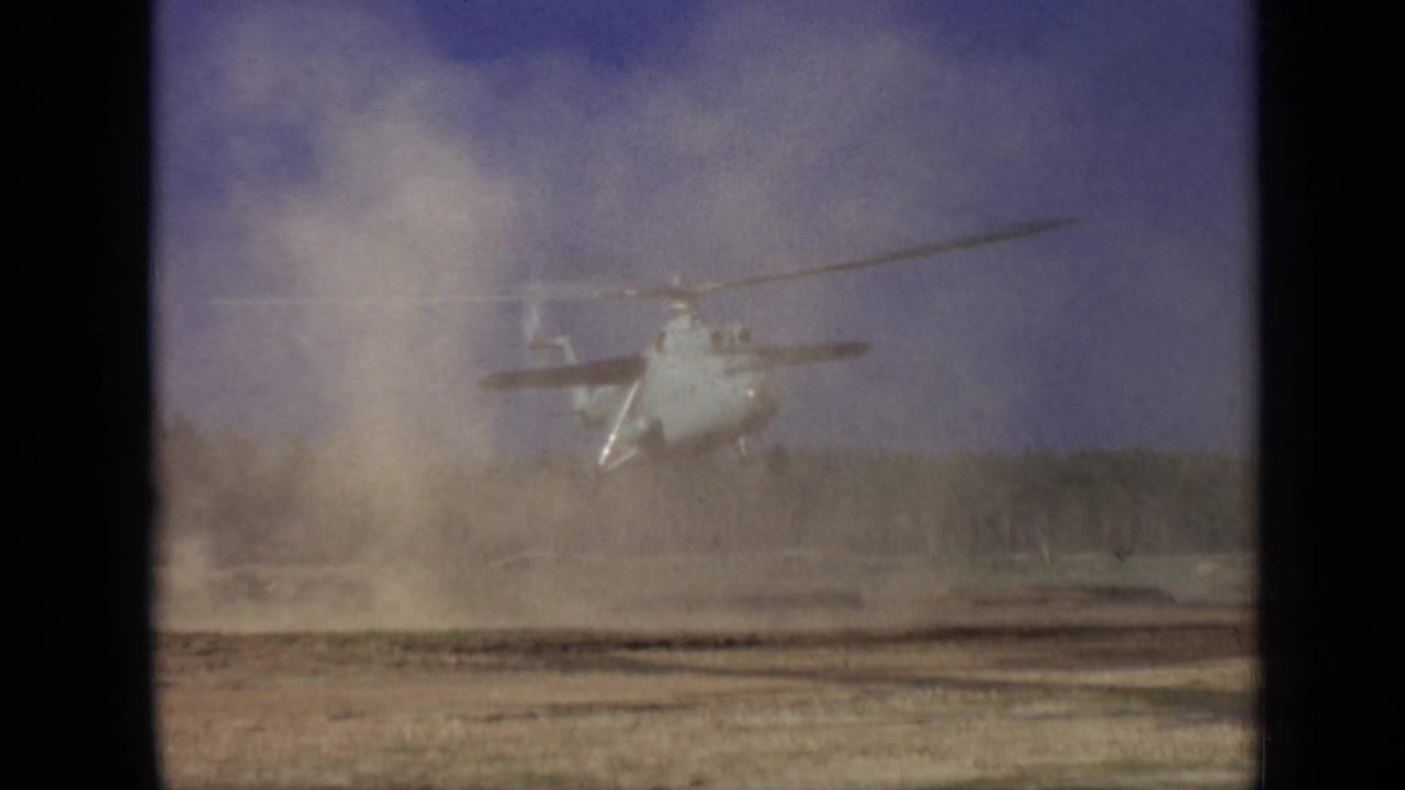 Helicopter hovering low over a dusty field with trees in the distance.