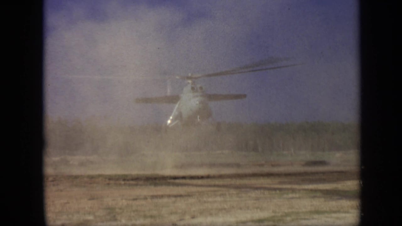Helicopter centered above a dusty field with rotor wash spreading across the ground.
