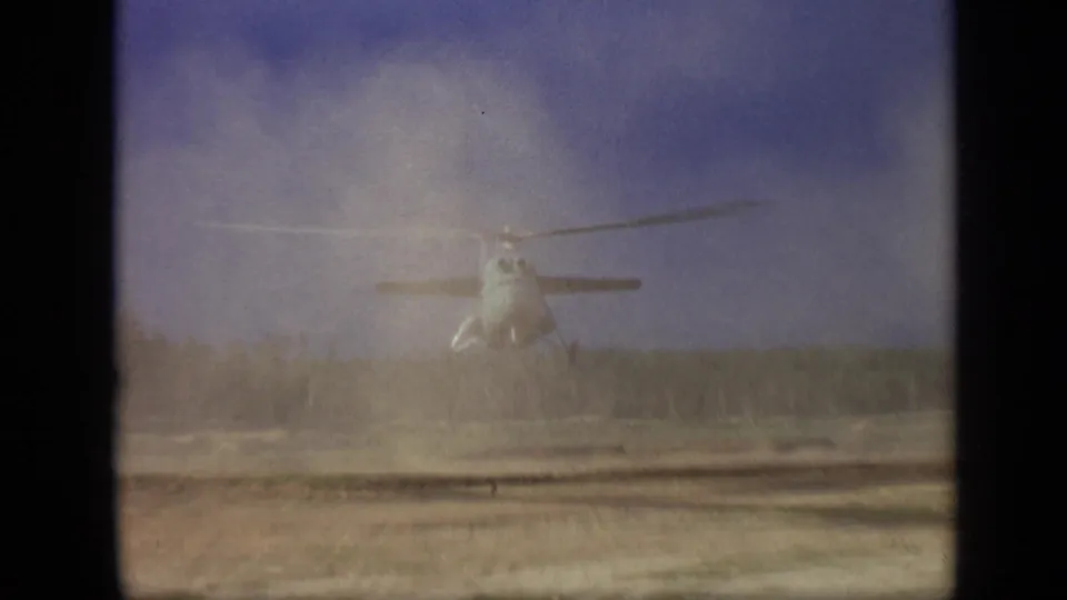 A 1971 8mm frame shows the helicopter holding a low hover while dust softens the view of the field and trees.