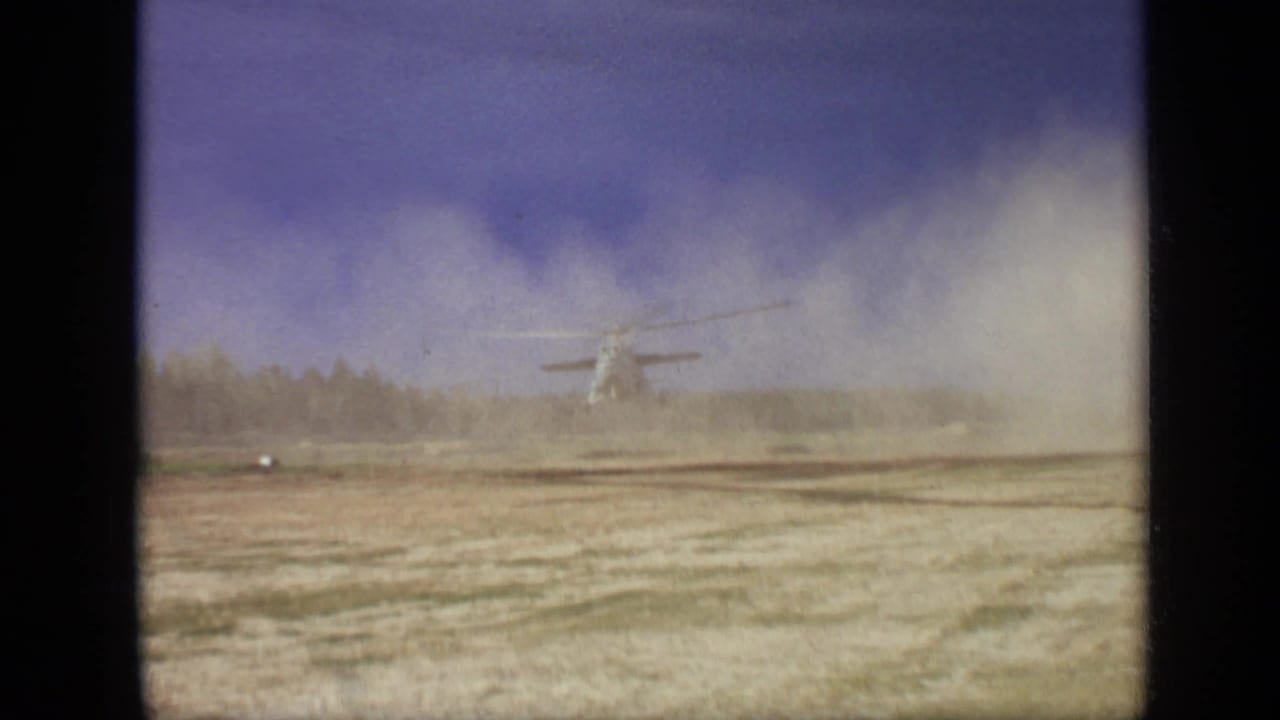 Distant helicopter hovering over a dusty open field with trees behind it.