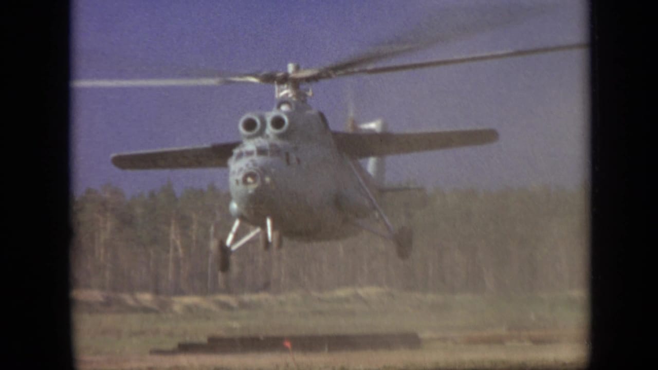 Large helicopter hovering low above a dirt field near a forest edge.