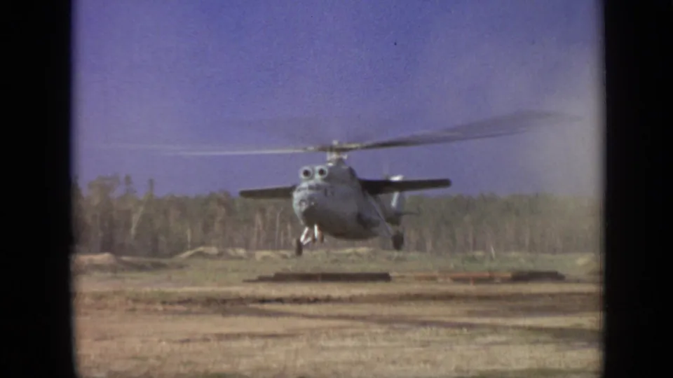 8mm frame of a large helicopter hovering over a rough field, with rotor wash and forested background visible.