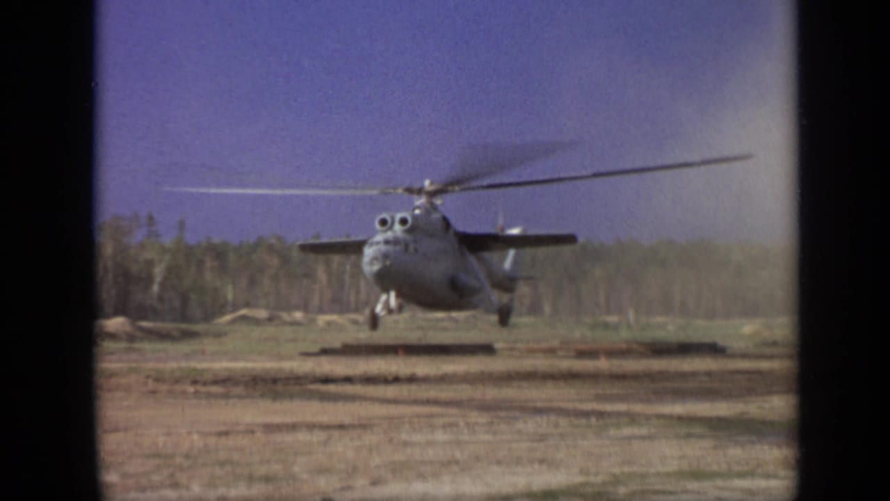 1971 Moscow-area 8mm frame showing a gray helicopter hovering low over a dirt field with rotor blur and a forest background.
