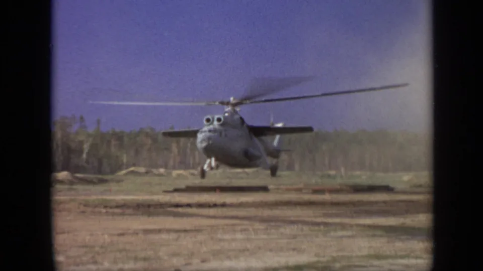 1971 Moscow-area 8mm frame showing a gray helicopter hovering low over a dirt field with rotor blur and a forest background.