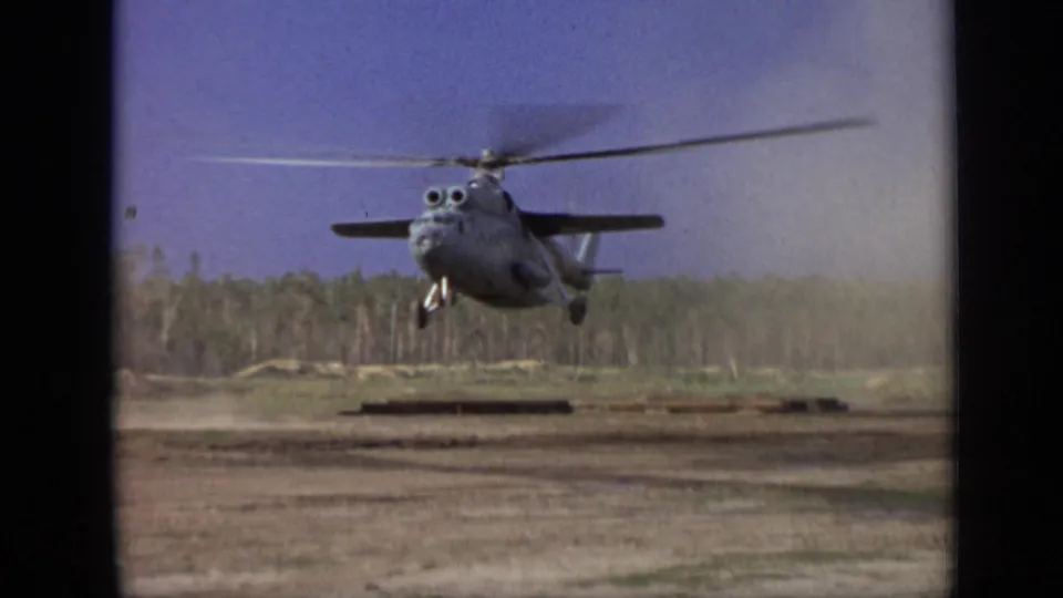 1971 Moscow-area 8mm frame of a gray helicopter hovering over a dirt field as dust drifts beside the aircraft.