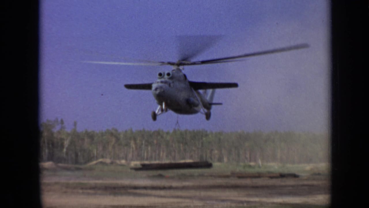 Large helicopter hovering over a clearing while carrying a long suspended beam.