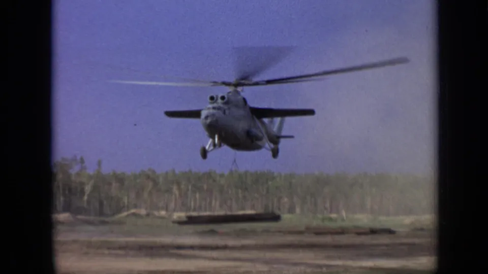 1971 Moscow-area 8mm frame showing a large gray helicopter lifting a long beam over open ground near a treeline.