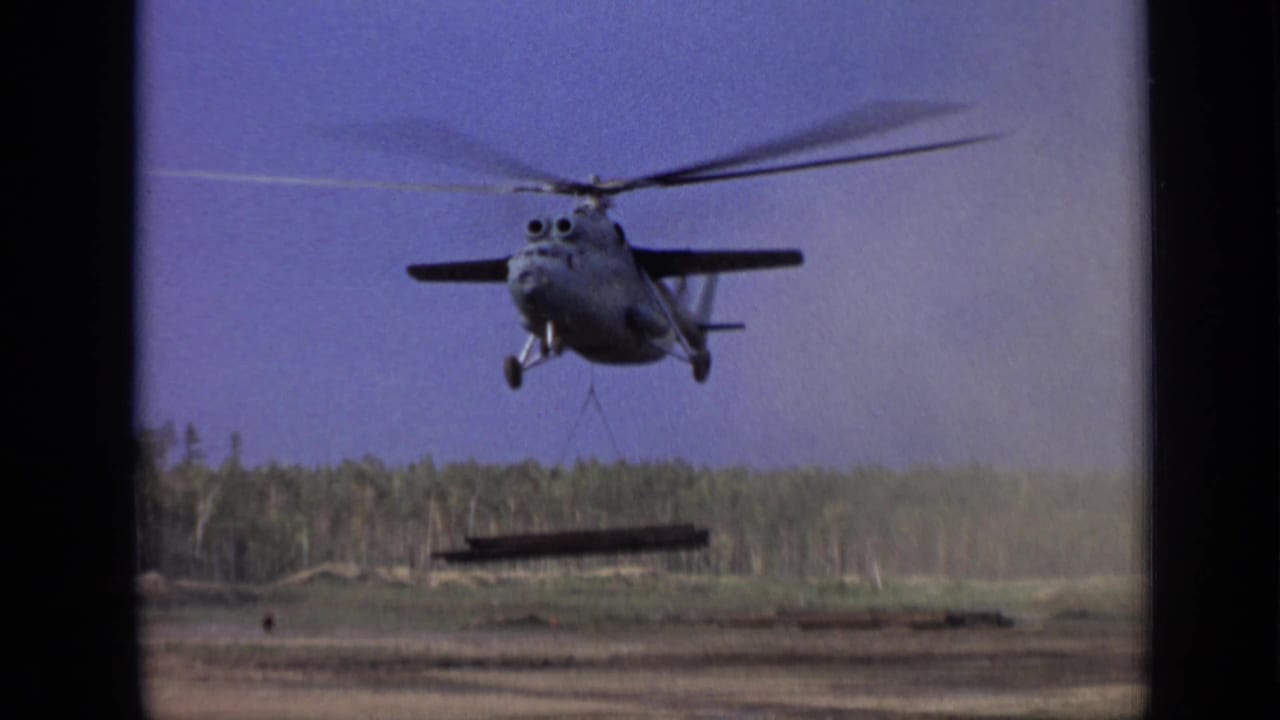 Helicopter facing the camera with a dark beam hanging below on cables.