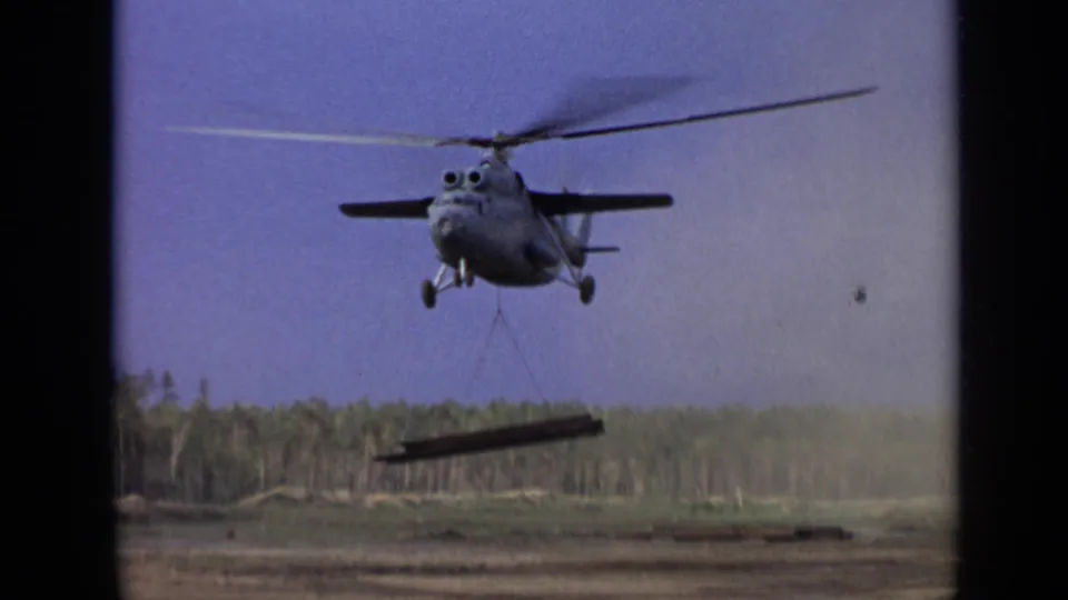 8mm frame of a large helicopter moving a suspended beam across a dirt clearing with forest in the background.