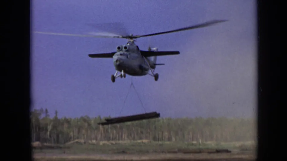 1971 8mm footage frame showing a large helicopter lifting a beam over open land near a forested area outside Moscow.