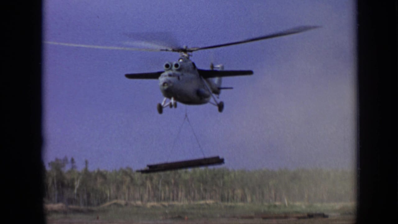 Helicopter lifting a long beam by sling over an open field and treeline.
