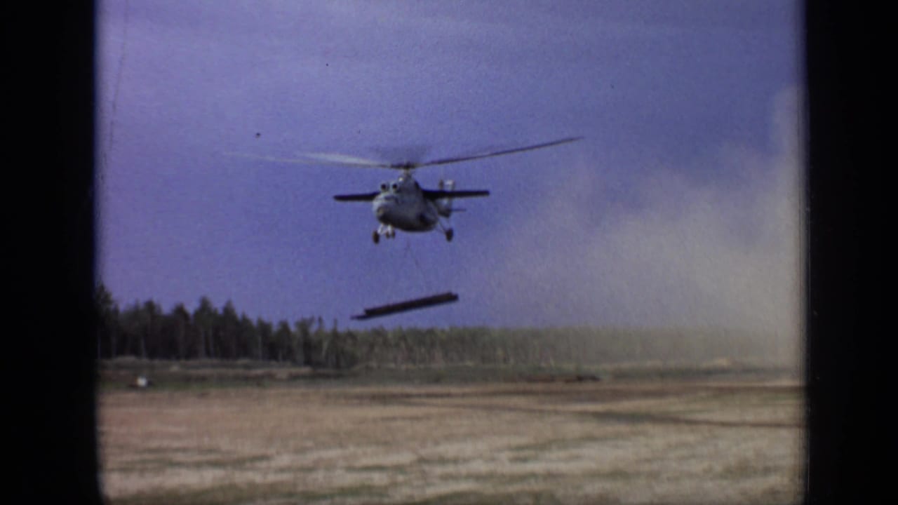 The helicopter moves across the Moscow-area field with a suspended beam below, framed by 8mm film borders and treeline.