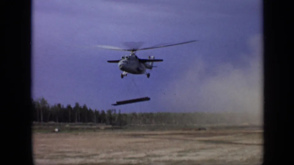 A Soviet-era helicopter, dated by metadata to 1971 Moscow, carries a beam across open terrain near a treeline.