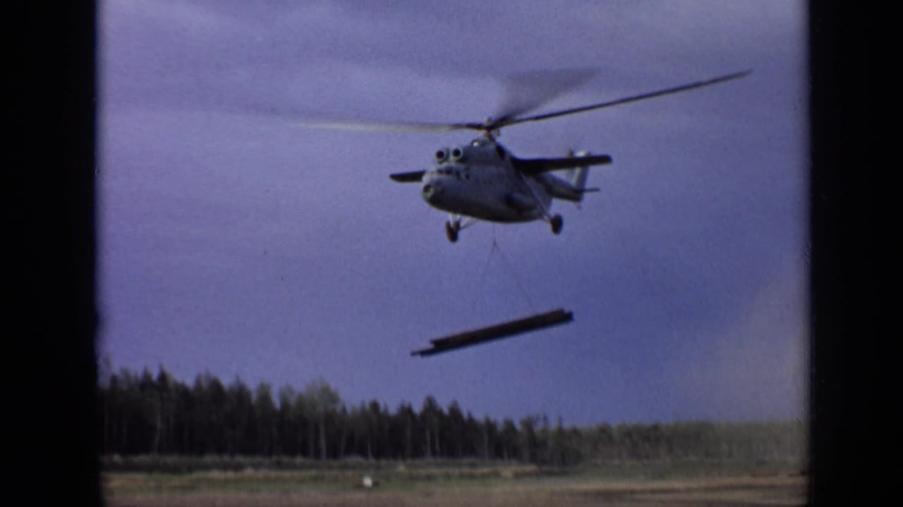 Helicopter carrying a suspended steel beam above open ground and a distant treeline.