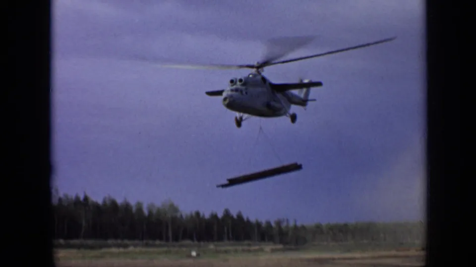 1971 Moscow 8mm frame showing a helicopter lifting a long steel beam over open land with a treeline below.