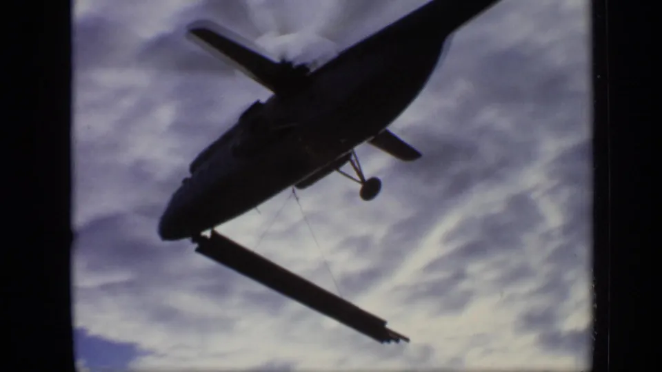Low-angle 1971 Moscow frame of a military-style helicopter silhouetted against clouds while carrying a steel beam.