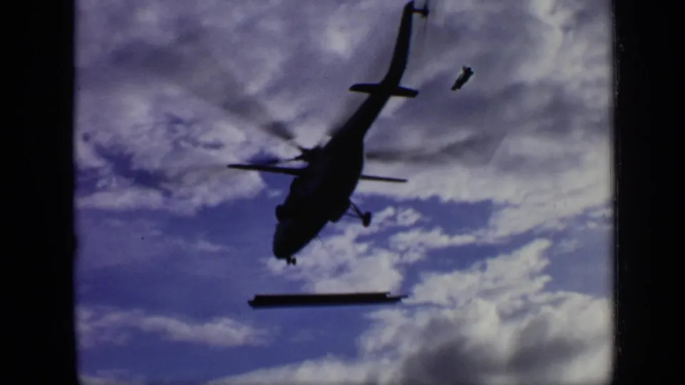 1971 Moscow 8mm frame looking upward at a helicopter carrying a steel beam under a cloudy sky.