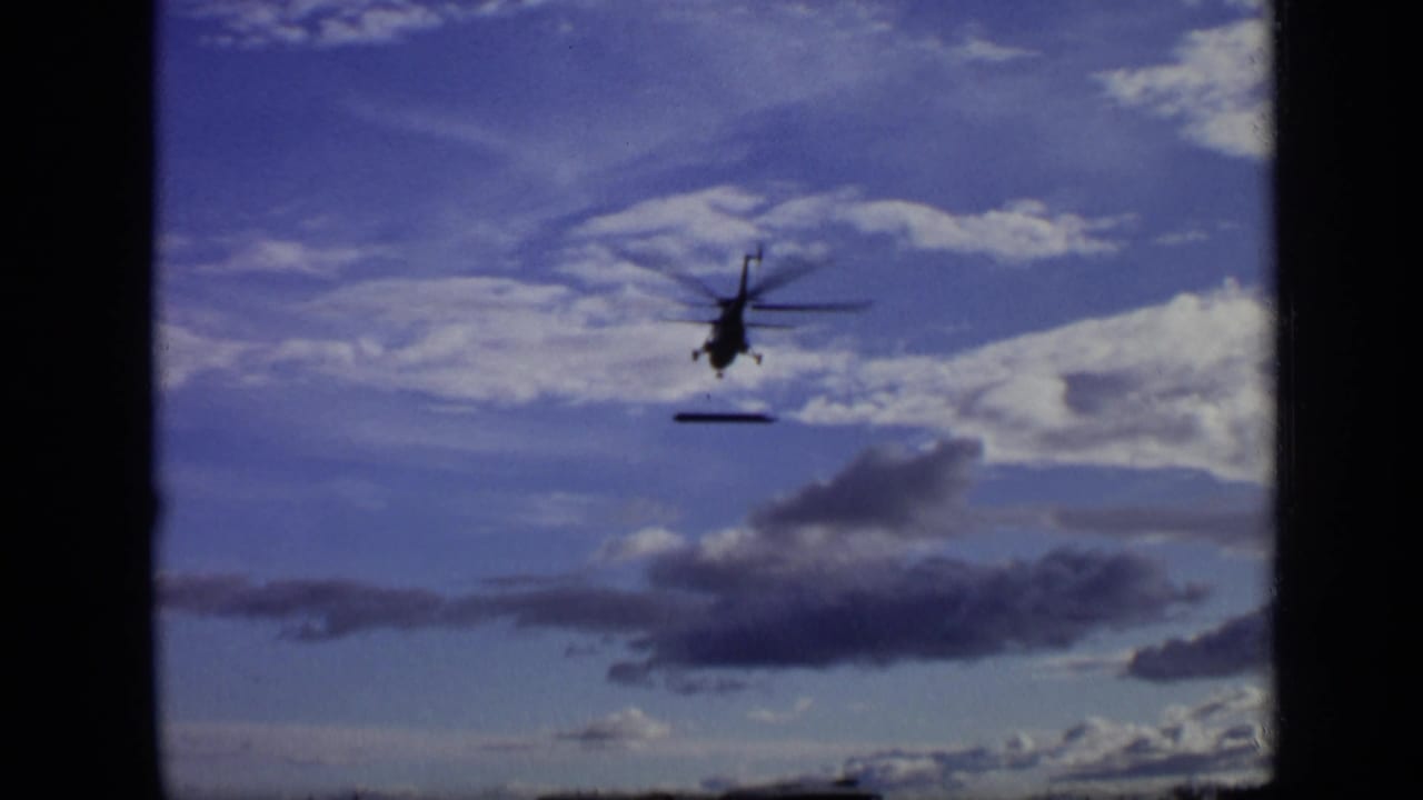 Silhouetted helicopter in a blue sky with clouds and a dark suspended load below.