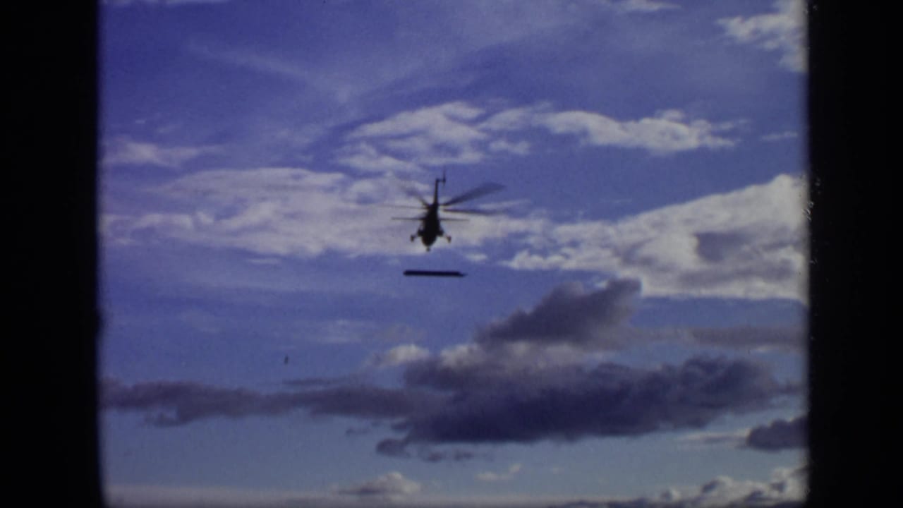 Distant helicopter against clouds with a narrow dark object hanging beneath it.