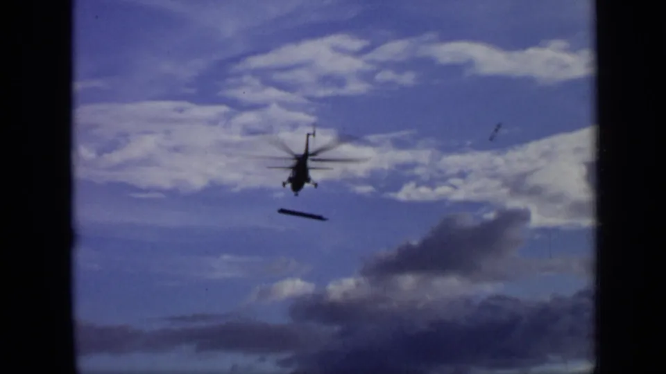 1971 Moscow 8mm frame showing the helicopter lower in the sky, silhouetted beneath bright cloud bands with a dark object below.