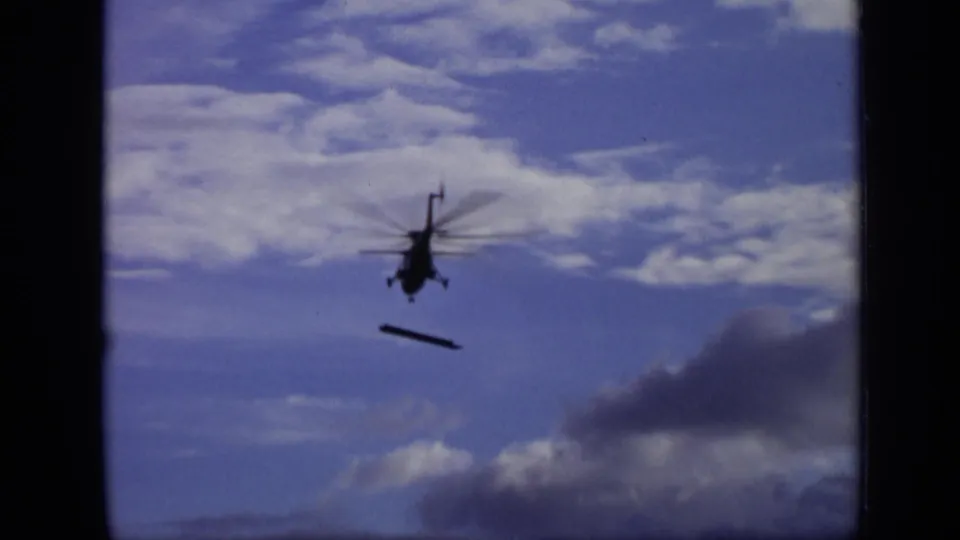 1971 Moscow 8mm frame of the helicopter continuing across the clouded blue sky with the suspended object angled below.