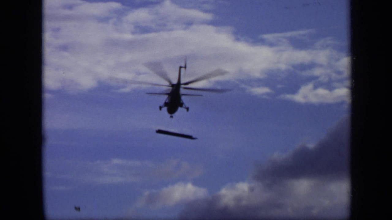 Helicopter silhouette carrying a long suspended object against a blue, clouded sky.
