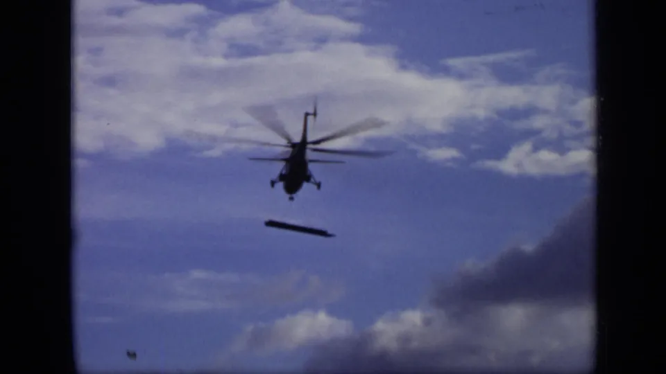 1971 Moscow 8mm frame showing a dark helicopter silhouette with a long bar-like load suspended beneath it against blue sky and clouds.