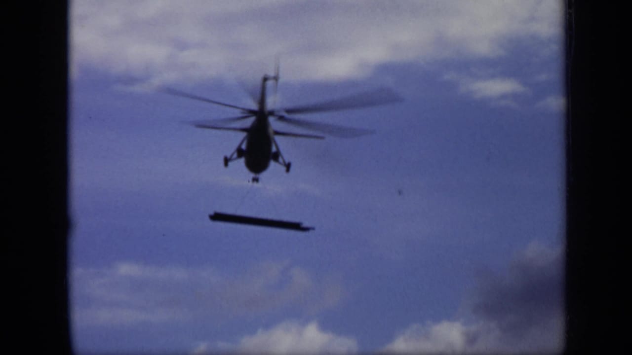 1971 Moscow 8mm frame with a head-on helicopter silhouette and a dark bar-like object hanging beneath it against clouded sky.