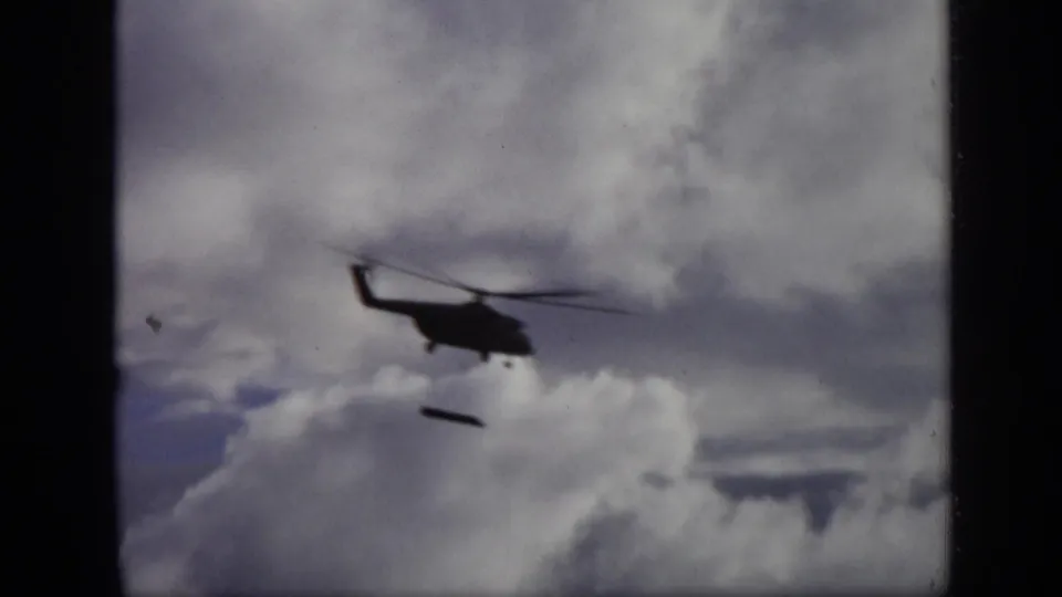 1971 Moscow 8mm frame showing the helicopter in profile with a small dark load below, partly silhouetted against gray and white clouds.