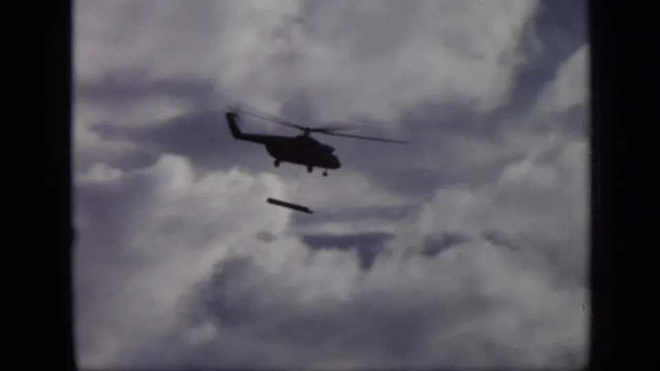 1971 Moscow 8mm frame showing a dark helicopter silhouette with a long object falling below it against thick clouds.
