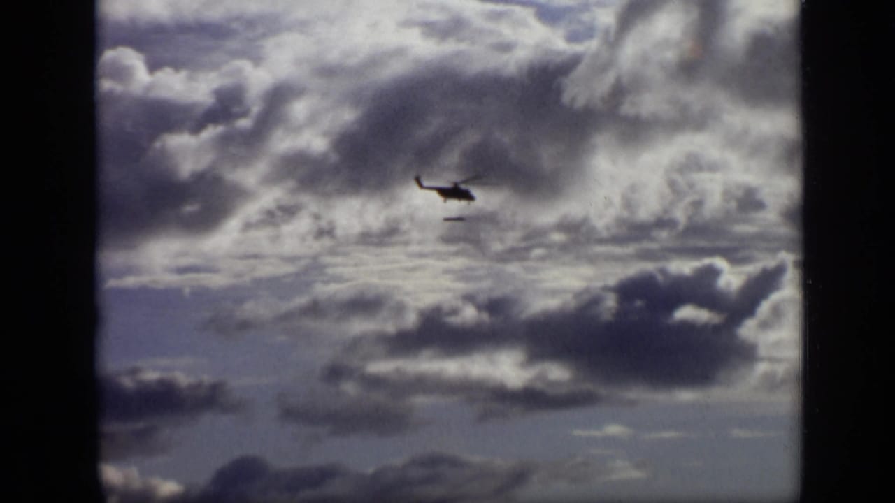 Helicopter silhouette flying beneath heavy clouds in a pale sky.