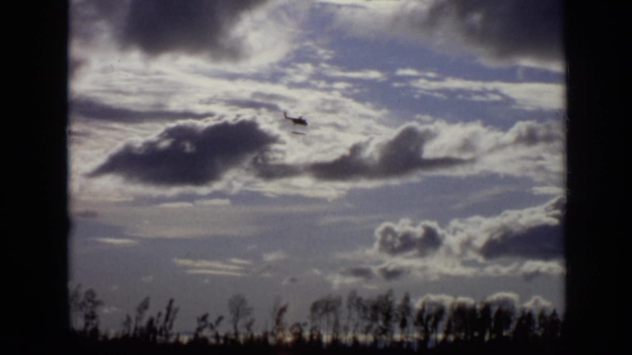 Small helicopter crossing a cloudy sky above a dark treeline.