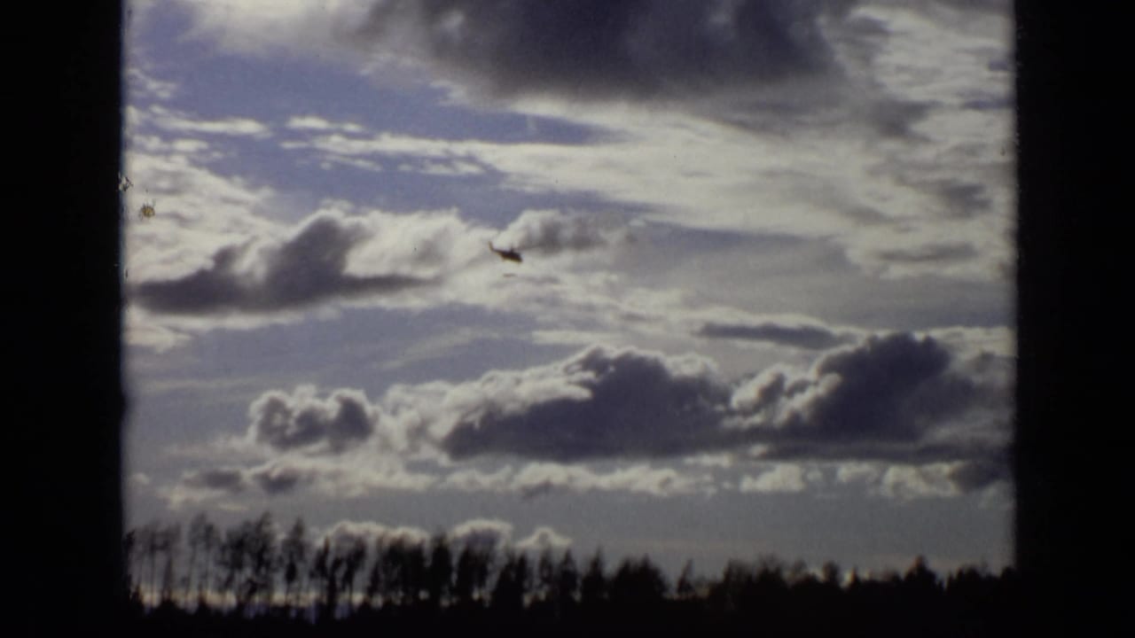 Small helicopter crossing a cloud-filled sky above a dark treeline.