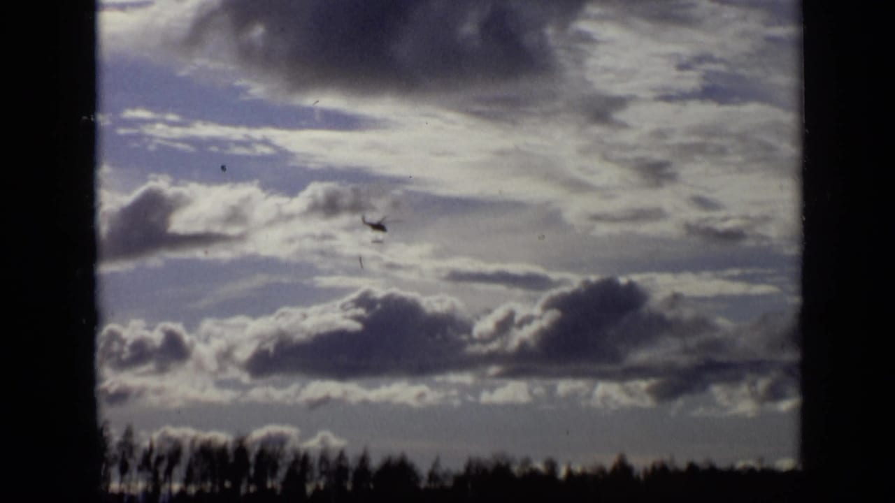 The helicopter remains visible in the cloudy sky above a low treeline, framed by dark 8mm gate edges.