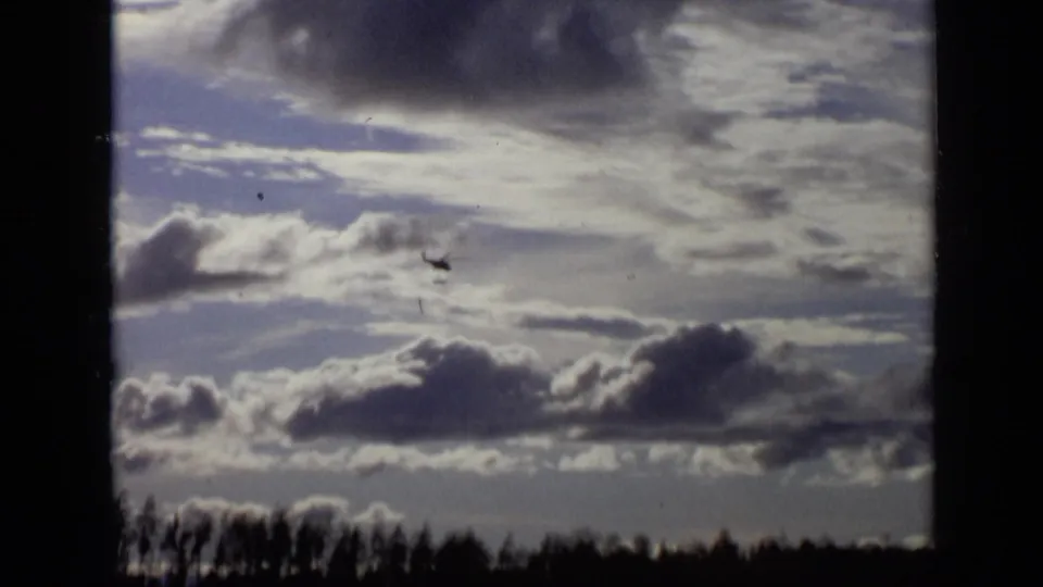 The helicopter remains visible in the cloudy sky above a low treeline, framed by dark 8mm gate edges.