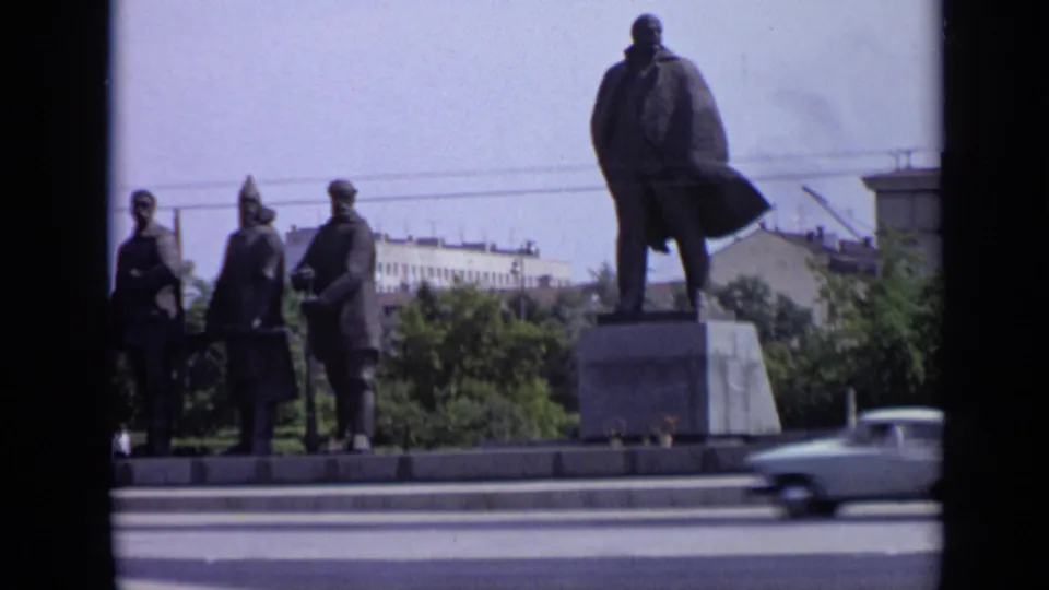 A Soviet-era monument in Moscow shows a large standing statue with soldier figures nearby as a car passes in front.