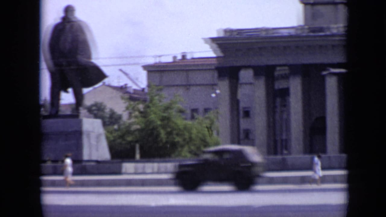 Statue, columned building, pedestrians, and a blurred car on a wide city street.